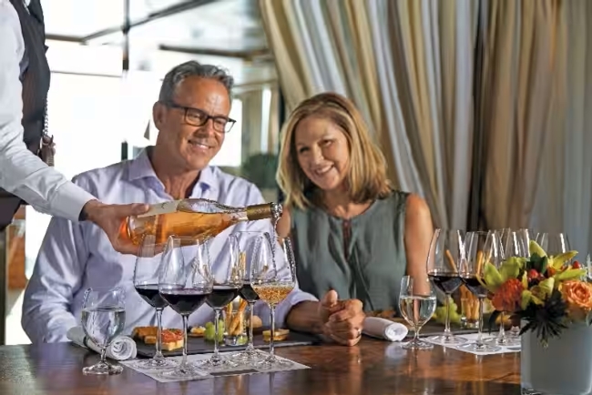 A sommelier pours rosé for a smiling couple seated at a wine tasting with flights of red and white wines