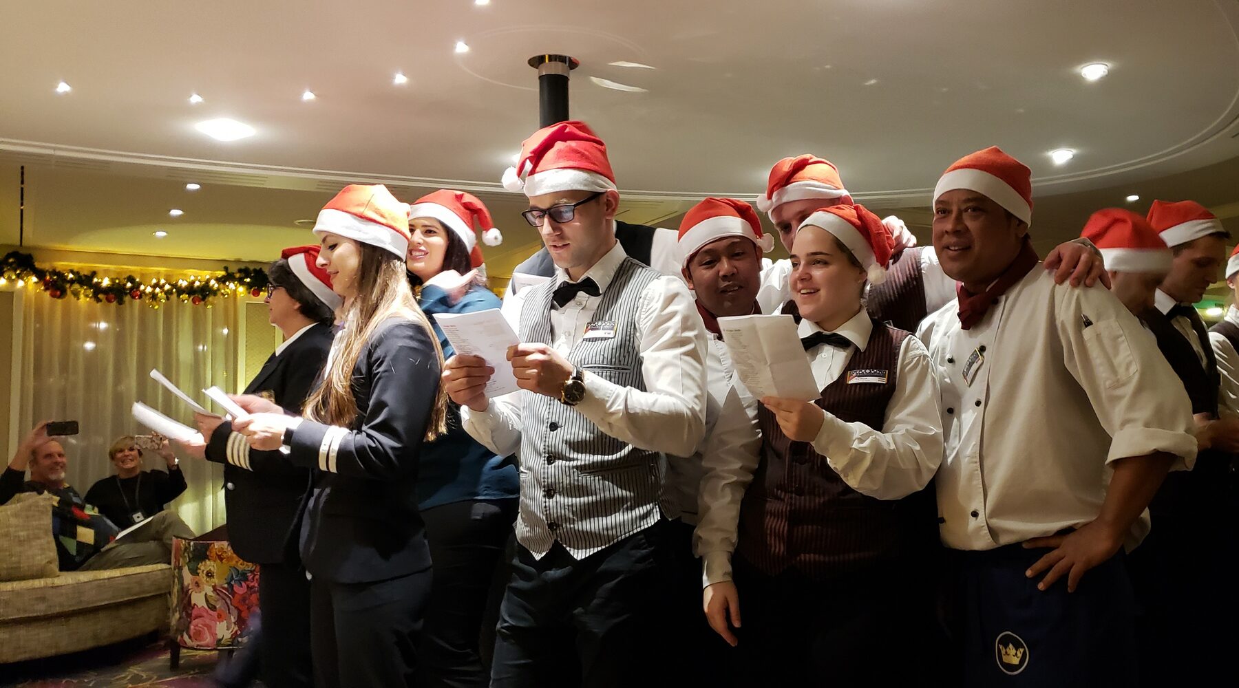 AmaStella crew members in Santa hats sing carols from song sheets in the ship's lounge
