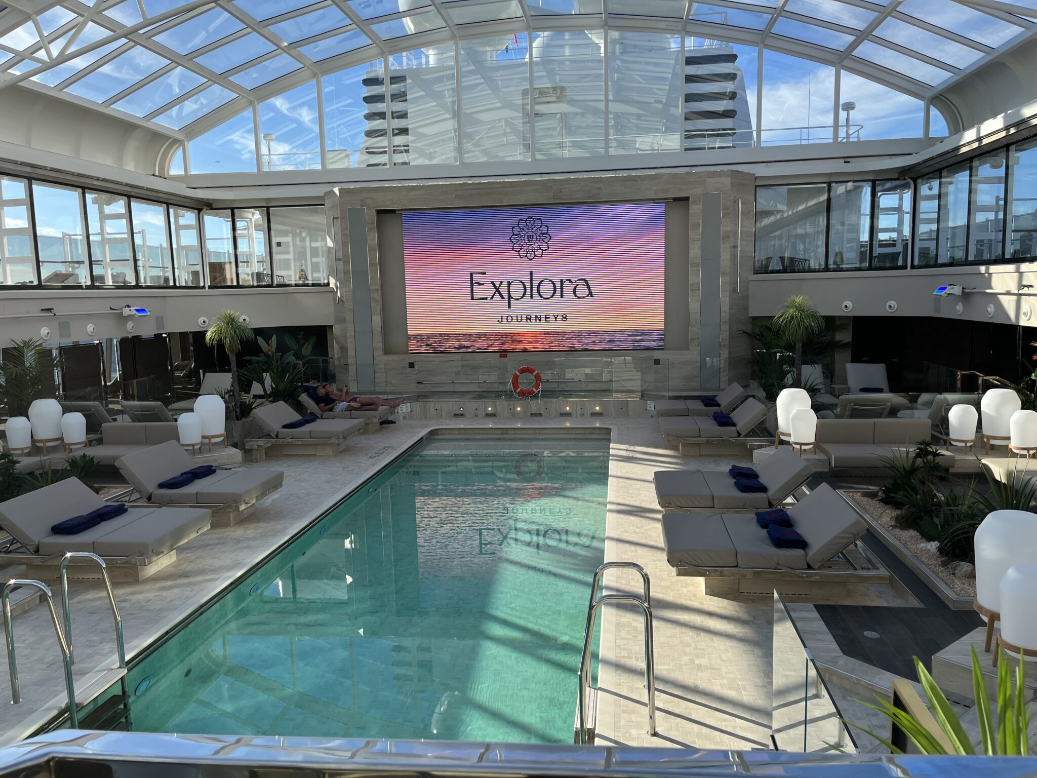 Conservatory Pool aboard Explora Journeys with retractable glass roof, lounge chairs, and the Explora Journeys brand screen