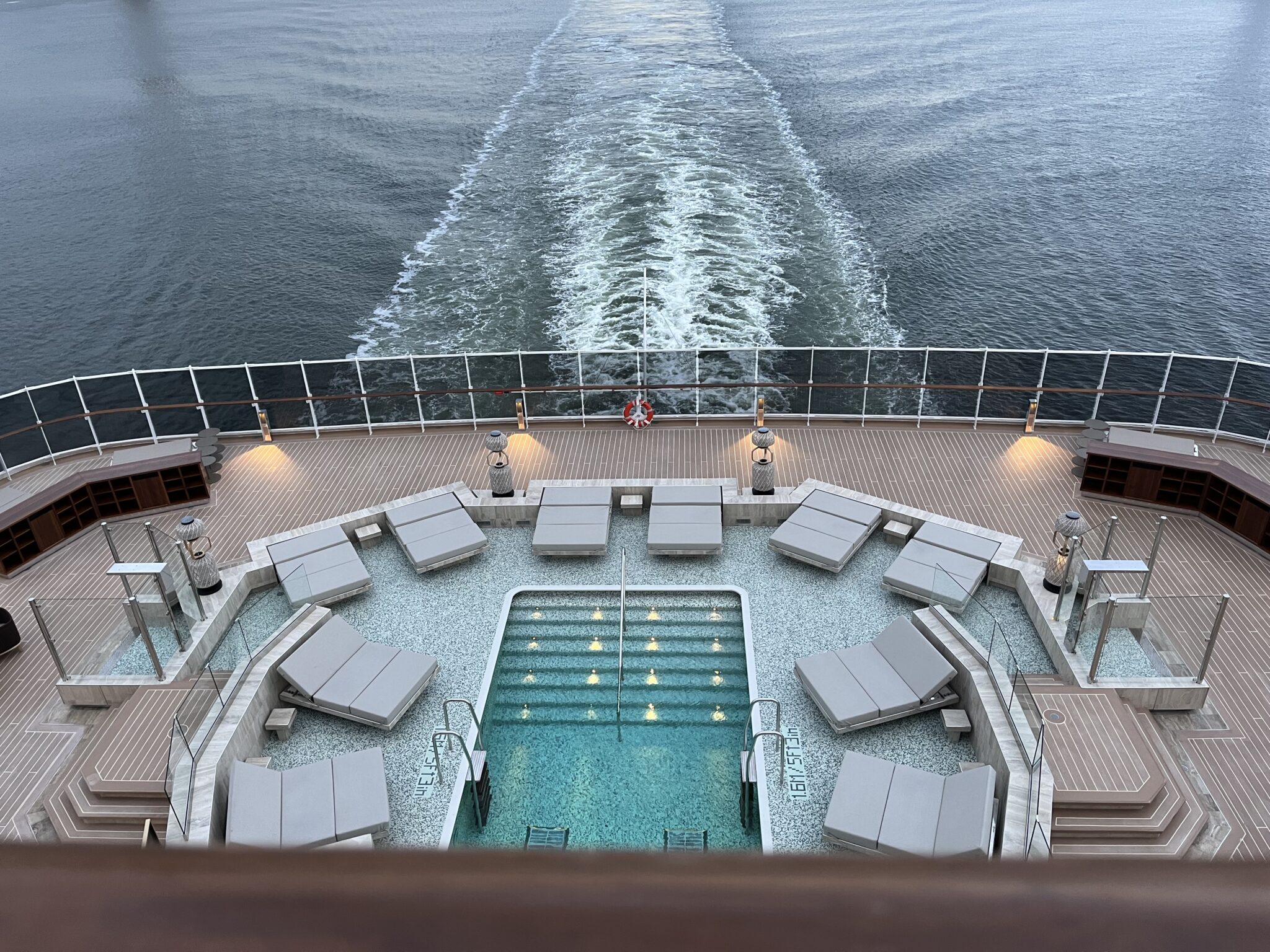 Aft pool deck at dusk aboard Explora Journeys with square pool, cushioned loungers, and ocean wake views
